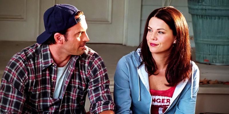 Luke And Lorelai looking at each other and smiling while sitting on the front steps of a house in Gilmore Girls, بهترین زوج های سریال درام