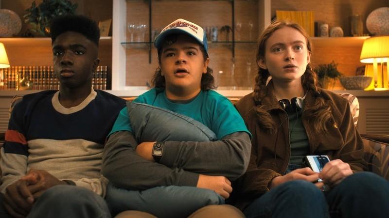 Lucas Dustin and Max sit together on a couch in Stranger Things Tales from '85