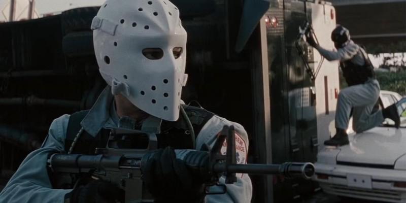 یک سارق با ماسک هاکی تفنگش را بالا گرفته در حالی که سارق دیگری در پس زمینه در Heat تلاش می کند وارد کامیون شود - دوئل Heat پاچینو دنیرو