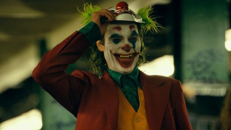 جوکر در حال خندیدن و بلند کردن یک ماسک دلقک در فیلم Joker، از بهترین فیلم های ابرقهرمانی پرفروش
